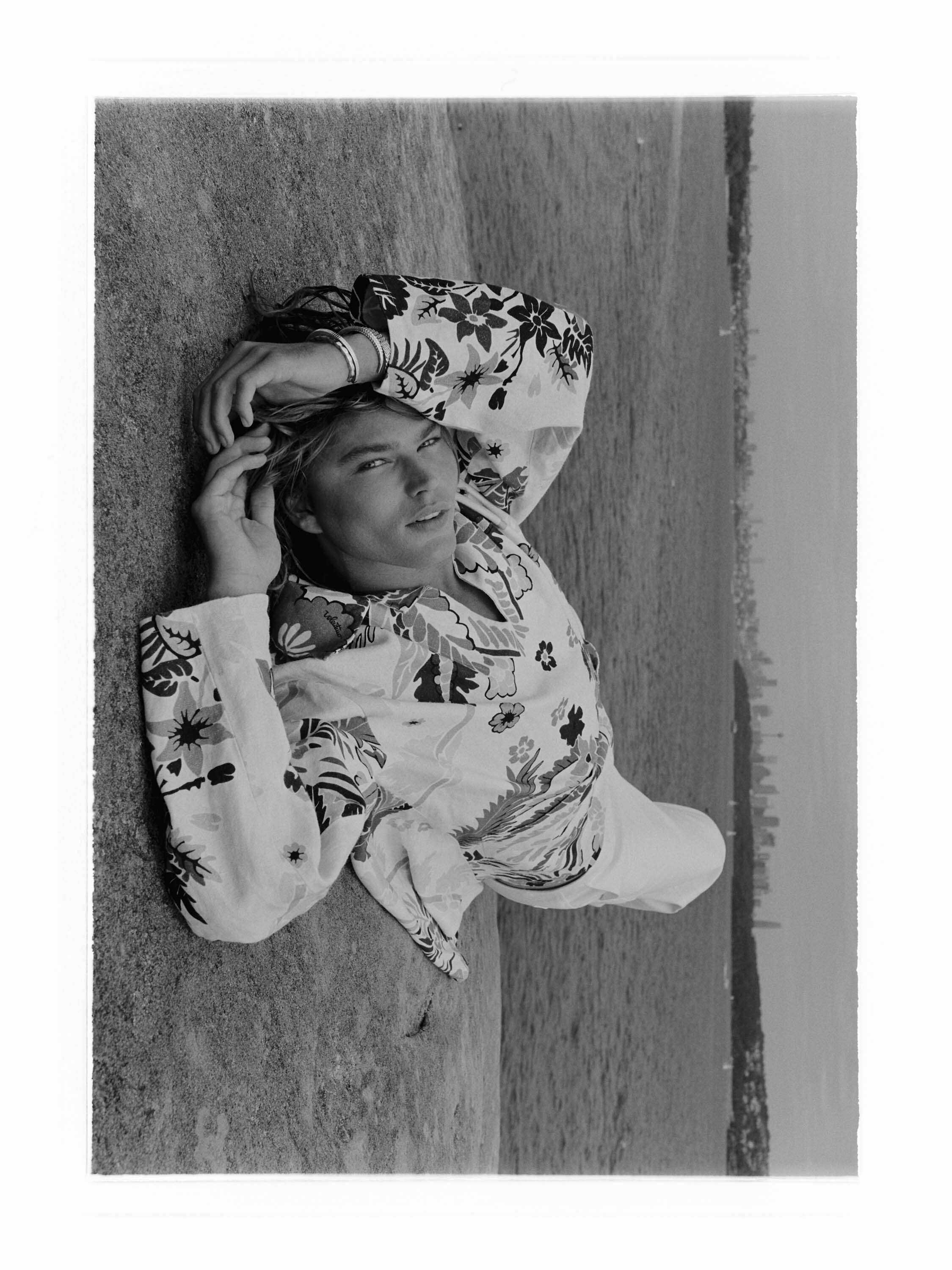 Black and white portrait of a young adult male in a floral shirt lying in a grassy field with a cityscape in the background.